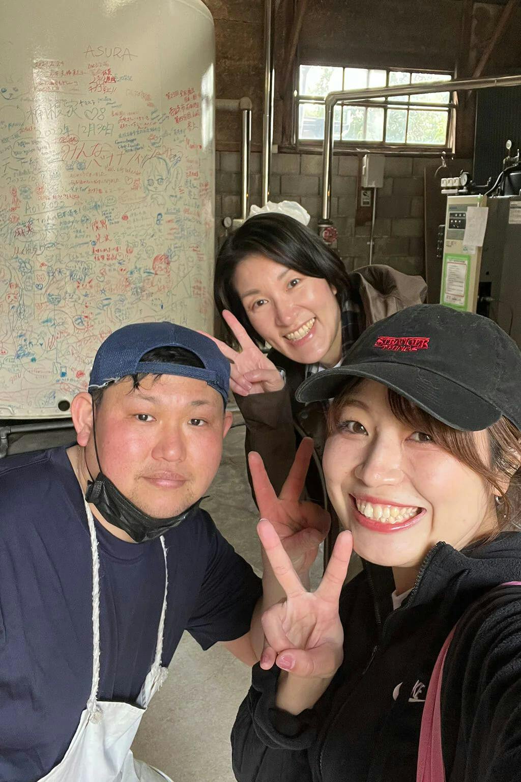 Mitsutake’s “kuraichi,” the toji’s second-in-command, poses for a picture with Azusa and Rina in front of a giant fermentation tank.